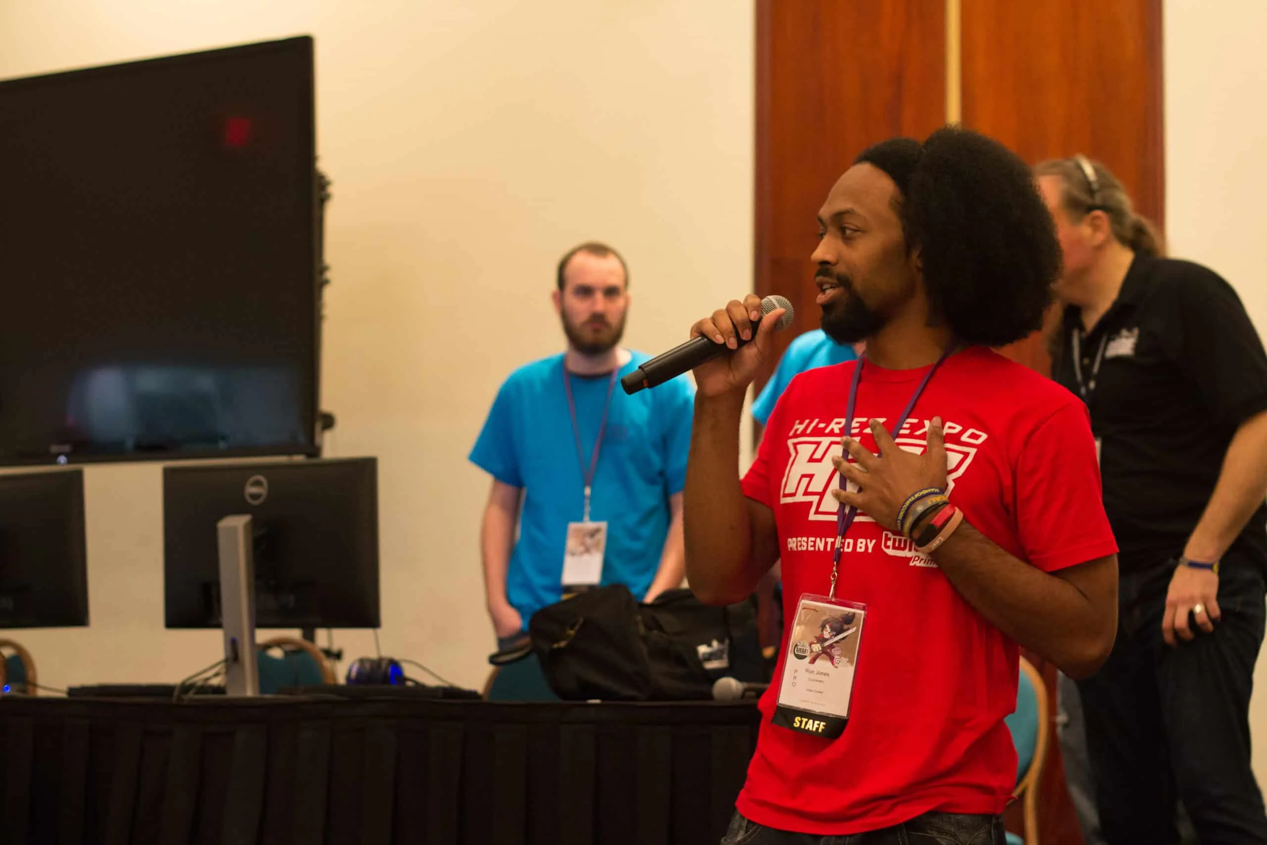 A person in a red shirt giving a speech or presentation, with an audience in the background at a conference or event.